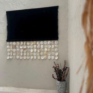 May include: A rectangular black fabric wall hanging featuring a bottom border of multiple rows of circular, iridescent white capiz shells. The shells are arranged in a grid pattern and are attached to the lower edge of the dark textile.