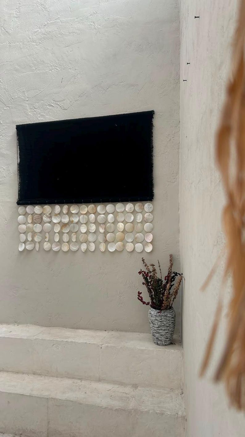 May include: A rectangular black fabric wall hanging featuring a bottom border of multiple rows of circular, iridescent white capiz shells. The shells are arranged in a grid pattern and are attached to the lower edge of the dark textile.