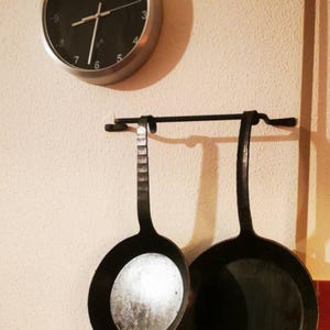 Op de afbeelding: Twee zwarte gietijzeren koekenpannen hangen aan een zwarte metalen rek dat aan een witte muur is bevestigd. De koekenpannen hebben lange handvatten en zijn klaar om te koken.