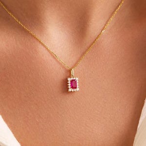 Könnte beinhalten: Eine goldfarbene Halskette mit einem rechteckigen, roten Edelsteinanhänger. Der Anhänger ist von kleinen, klaren Steinen umgeben. Die Kette wird auf einem weißen Hemd getragen.