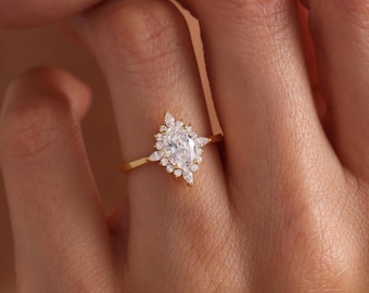 May include: A gold ring featuring a central marquise-cut clear gemstone surrounded by a halo of smaller round and marquise-cut stones. The band is thin and polished gold, creating a delicate look with a cluster of sparkling white stones.