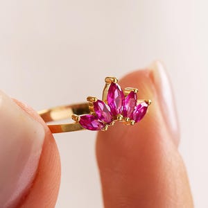 Peut inclure: Une bague en or ornée d'un groupe de pierres précieuses roses vif taillées en marquise. Le corps de la bague est fin et poli, et les pierres sont disposées en éventail. La bague est tenue entre deux doigts.