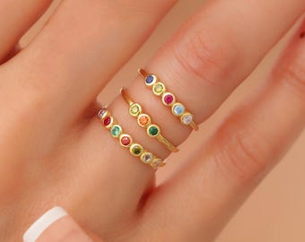 May include: Three gold rings, each featuring a row of small, round, multi-colored gemstones. The rings are worn on a finger, showcasing a variety of hues including red, green, yellow, orange, blue, and clear stones set in thin gold bands.