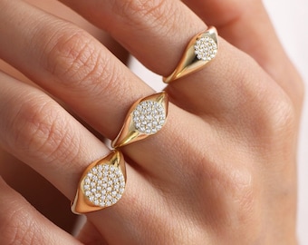 May include: Three gold-colored rings with a teardrop shape, each featuring a circular cluster of small, sparkling stones. The rings are displayed on a person's fingers, showcasing their design and shine. The background is a soft, neutral color.