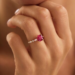 Könnte beinhalten: Ein Goldring mit einem großen, kissenförmigen, leuchtend pink-roten Edelstein als Mittelstück. Die Ringschiene ist mit einer Reihe kleinerer, klarer, marquise- und rundgeschliffener Steine verziert, die sich entlang des Goldbandes erstrecken.