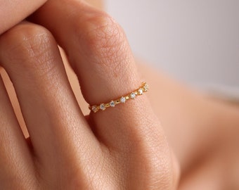 May include: A delicate gold ring with small, evenly spaced clear gemstones. The ring is worn on a finger, with the hand and part of the face visible. The ring's design is simple and elegant, suitable for everyday wear.