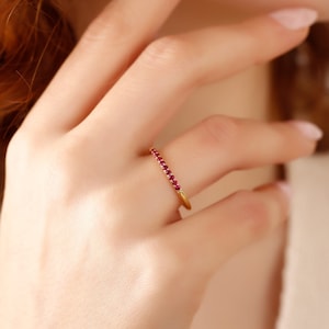 Könnte beinhalten: Ein zarter Goldring mit einer Reihe kleiner, roter Edelsteine. Der Ring wird am Finger einer Person mit roten Haaren getragen.