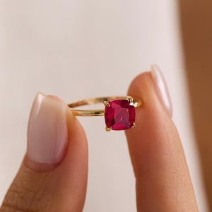 May include: A gold ring featuring a cushion-cut, vibrant red gemstone set in a four-prong setting. The band is thin and polished, showcasing a smooth, metallic finish that complements the deep red colour of the central stone held in place.