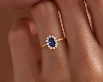 May include: A gold ring featuring a central oval-cut deep blue sapphire surrounded by a halo of twelve small, round, clear white stones. The thin, polished gold band supports the floral-shaped setting, creating a delicate and refined appearance.