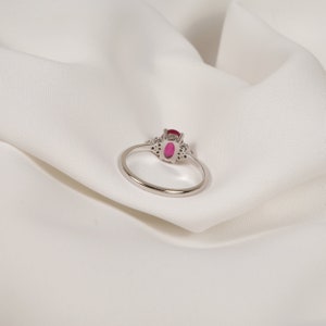 Puede incluir: Un anillo de plata con una gran piedra de rub&iacute; ovalada rodeada de diamantes m&aacute;s peque&ntilde;os. El anillo est&aacute; sobre un fondo de tela blanca.