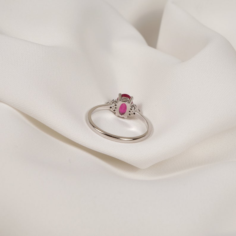 Puede incluir: Un anillo de plata con una gran piedra de rub&iacute; ovalada rodeada de diamantes m&aacute;s peque&ntilde;os. El anillo est&aacute; sobre un fondo de tela blanca.