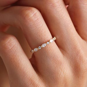 Könnte beinhalten: Ein dünner Goldring mit einem abwechselnden Muster aus marquise- und rundgeschliffenen klaren Steinen. Die Steine sind entlang der oberen Hälfte des zarten Metallbandes gefasst und erzeugen ein durchgehendes, funkelndes Design am Finger.