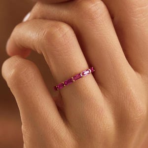 May include: A thin gold band ring featuring a series of vibrant, rectangular-cut pink gemstones set in a row. The stones are arranged horizontally along the band, held in place by small gold prongs, creating a continuous, elegant, and colorful design.