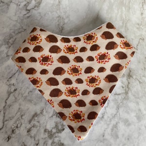 May include: A beige bandana with a white border features a repeating pattern of brown hedgehogs surrounded by orange and red leaves.