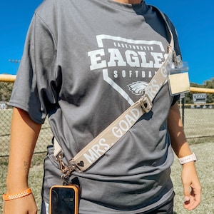 May include: A gray t-shirt with the text "East Forsyth Eagles Softball" printed on the front. A beige phone strap with the text "Mrs. Goad" is worn diagonally across the chest.