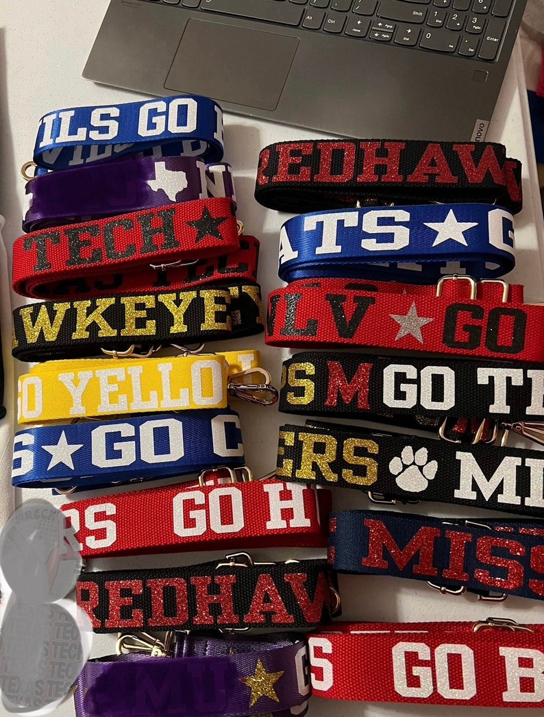 以下が含まれることがあります： 「ILS GO」、「REDHAW」、「TS」、「WKEYEY」、「LV GO」、「S M GO TI」、「ERS」、「S GO H」、「REDHAW」、「MISS」、「S GO B」など、さまざまな大学チームの名前とロゴが書かれたカラフルなファブリックストラップのスタック。