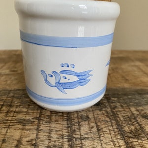 Può includere: Un barattolo di ceramica bianca con un tappo di sughero. Il barattolo ha una striscia blu al centro e un disegno di pesce blu. Il barattolo è appoggiato su una superficie di legno.