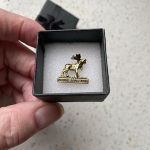 May include: A gold-toned metal lapel pin shaped like a standing moose, featuring the text MOOSE JAW SASK embossed on the base. The pin is presented inside a small, square black gift box with a white foam insert for secure placement and display.