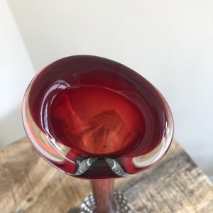 Peut inclure: Un vase en verre rouge avec un dessus évasé et une longue tige élancée. Le vase a une base bulbeuse avec de petits accents ronds et transparents. Le verre est d'un rouge profond et riche, et la tige semble être brun foncé.