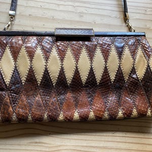 May include: A vintage handbag featuring a diamond pattern in shades of brown, tan, and cream. It has a dark brown frame and a matching shoulder strap. The clasp is rectangular with a wooden detail.