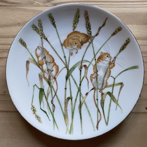 May include: White ceramic plate featuring a detailed illustration of three small, brown mice amongst wheat stalks. The plate has a thin, dark blue rim. The artwork uses green and tan colours. The plate is set on a wooden surface.