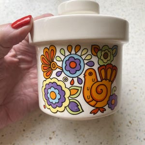 Può includere: Un barattolo in ceramica color crema con coperchio abbinato, caratterizzato da una stampa grafica colorata di un uccello arancione stilizzato circondato da vari fiori nelle tonalità del viola, giallo, arancione e verde. Il contenitore è cilindrico.