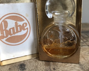 Puede incluir: Frasco de perfume vintage "Babe" en un estuche dorado. El frasco de vidrio transparente contiene un líquido dorado y tiene un tapón transparente y redondeado. El estuche está abierto, revelando una tarjeta con la palabra "Babe" en un diseño circular.