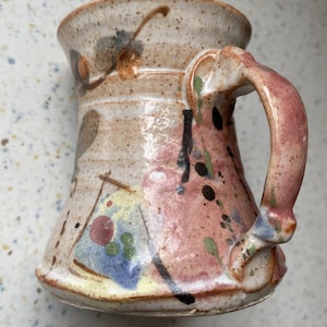 Könnte beinhalten: Ein Keramikbecher mit gesprenkelter beiger Basis und abstrakten, handgemalten Mustern in Pink, Blau, Gelb und Schwarz. Der Becher hat einen ausgestellten Rand, einen geschwungenen Henkel und eine glänzende Oberfläche mit Farbspritzern.