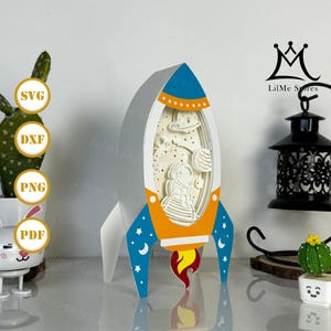 May include: A layered paper art rocket ship in shades of white, blue, orange, and red. The rocket features a detailed interior with an astronaut, planets, and stars. The design includes the text "SVG", "DXF", "PNG", and "PDF".