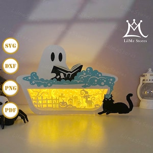 Könnte beinhalten: Eine beleuchtete Schattenbox zum Thema Halloween. Sie zeigt einen Geist, der in einer Badewanne mit blauen Blasen liest, und eine schwarze Katze mit Hexenhut. Die Box hat eine von innen beleuchtete Friedhofsszene.