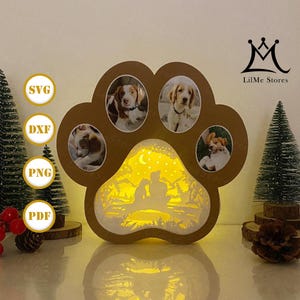 Könnte beinhalten: Ein brauner, pfotenförmiger Fotorahmen mit vier Hundefotos und einer Hintergrundbeleuchtungsszene eines Paares und eines Hundes unter einem Halbmond. Der Rahmen trägt die Texte "SVG", "DXF", "PNG" und "PDF". Das LilMe Stores-Logo ist sichtbar.