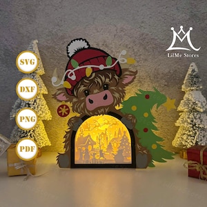 May include: A layered paper Christmas decoration featuring a cartoon Highland cow wearing a red hat and string lights. The cow holds a Christmas tree and a decorative archway with a lit winter village scene. The image includes the text "SVG", "DXF", "PNG", and "PDF".