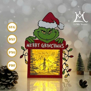 Puede incluir: Decoración navideña de papel en capas con el Grinch con un gorro de Papá Noel, sosteniendo un letrero que dice "MERRY GRINCHMAS". La escena interior muestra una silueta del Grinch y Villa Quien. Luces decorativas y pequeños árboles también están incluidos.