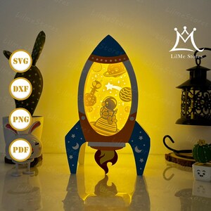 May include: A blue, orange, and yellow rocket-shaped night light with an astronaut and planets inside. The rocket has a star and moon design on the fins. The light is from LilMe Stores.
