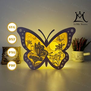 Puede incluir: Una lámpara decorativa con forma de mariposa, con un marco morado y un interior amarillo. El diseño presenta mariposas, flores y una luna recortadas. La luz está encendida, creando un brillo cálido. El texto "LilMe Stores" es visible.