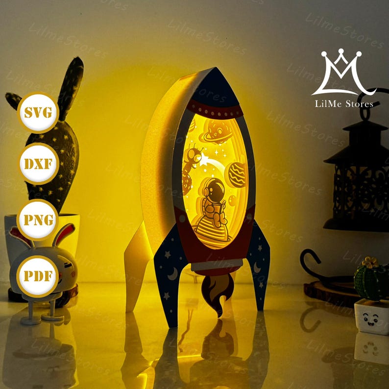 May include: A layered paper art rocket ship illuminated from within, featuring an astronaut reading in space. The rocket has red, white, and blue accents. The image also includes a cactus, a bunny figurine, and the text "SVG, DXF, PNG, PDF".