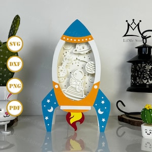 May include: A layered paper art rocket ship in blue, orange, and white, featuring an astronaut, planets, and stars. The rocket has a flame design at the bottom. The image also includes the text "SVG", "DXF", "PNG", and "PDF".