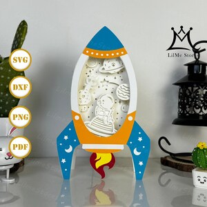 May include: A layered paper art rocket ship in blue, orange, and white, featuring an astronaut, planets, and stars. The rocket has a flame design at the bottom. The image also includes the text "SVG", "DXF", "PNG", and "PDF".
