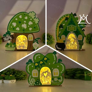 Puede incluir: Tres adornos decorativos iluminados con el tema del Día de San Patricio. Los adornos son verdes y marrones, con tréboles, gnomos y las palabras "Lucky" y "Good Luck". Los adornos están iluminados desde dentro, creando un brillo cálido.