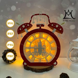 May include: A red and white layered paper clock with a castle design illuminated from within. The clock features reindeer antlers and stars, with the text "SVG", "DXF", "PNG", and "PDF" on the left side. The clock sits on a wooden base.