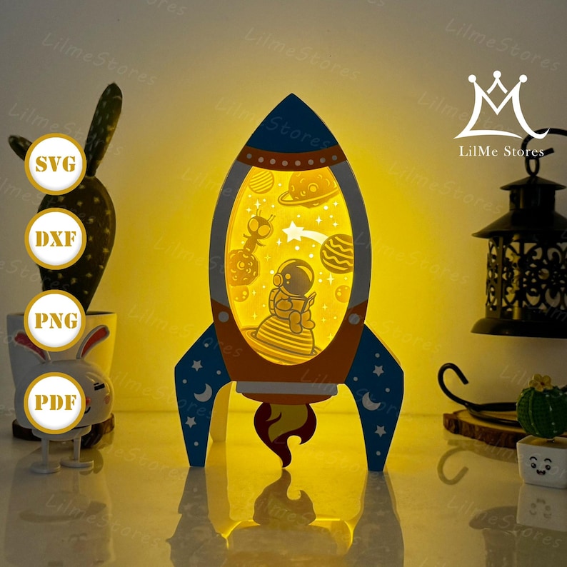 May include: A blue, orange, and yellow rocket-shaped night light with an astronaut and planets inside. The rocket has a star and moon design on the fins. The light is from LilMe Stores.