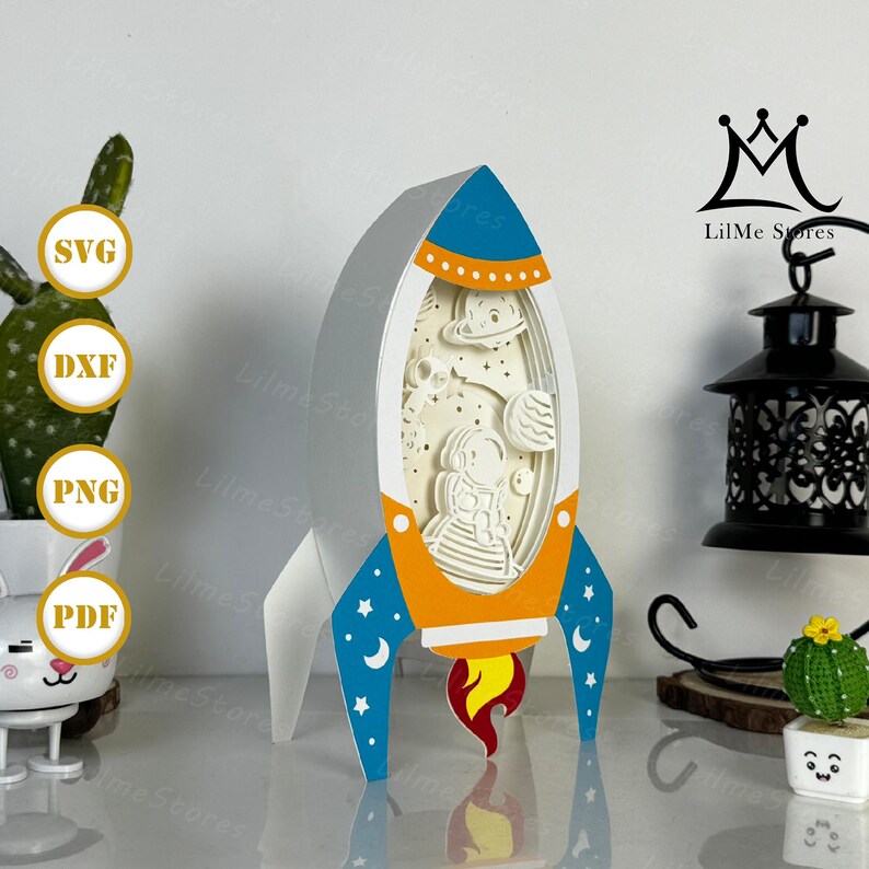 May include: A layered paper art rocket ship in shades of white, blue, orange, and red. The rocket features a detailed interior with an astronaut, planets, and stars. The design includes the text "SVG", "DXF", "PNG", and "PDF".