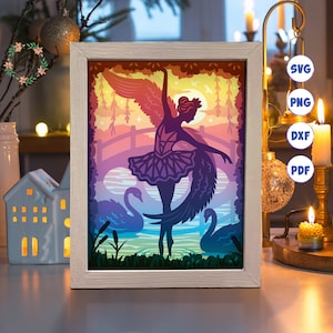 Könnte beinhalten: Ein mehrschichtiges Papier-Schnittbild einer Ballerina, die mit einem Schwan in einer Waldlandschaft tanzt. Die Ballerina trägt einen Tutu und Flügel. Das Design ist in einem weißen Rahmen gerahmt.