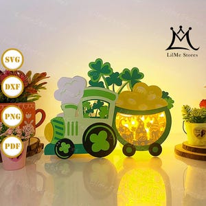 May include: A green and white St. Patrick's Day themed train with a pot of gold car. The train features shamrocks and a pot of gold filled with gnomes. The train is illuminated from within, creating a warm glow.