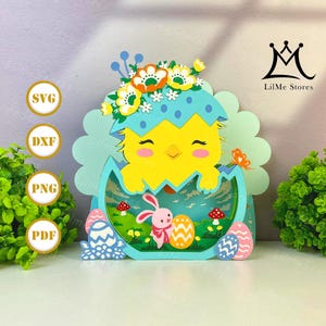 Puede incluir: Decoración de Pascua de papel en capas con un pollito amarillo saliendo de una cáscara de huevo azul, junto con un conejito rosa y huevos decorados. El diseño incluye flores y los textos "SVG", "DXF", "PNG" y "PDF". El logotipo de LilMe Stores es visible.