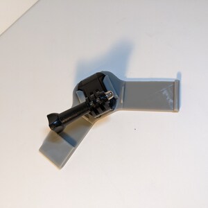 Pu&ograve; includere: Un supporto per fotocamera in plastica grigia con un supporto per fotocamera in stile GoPro nero attaccato.