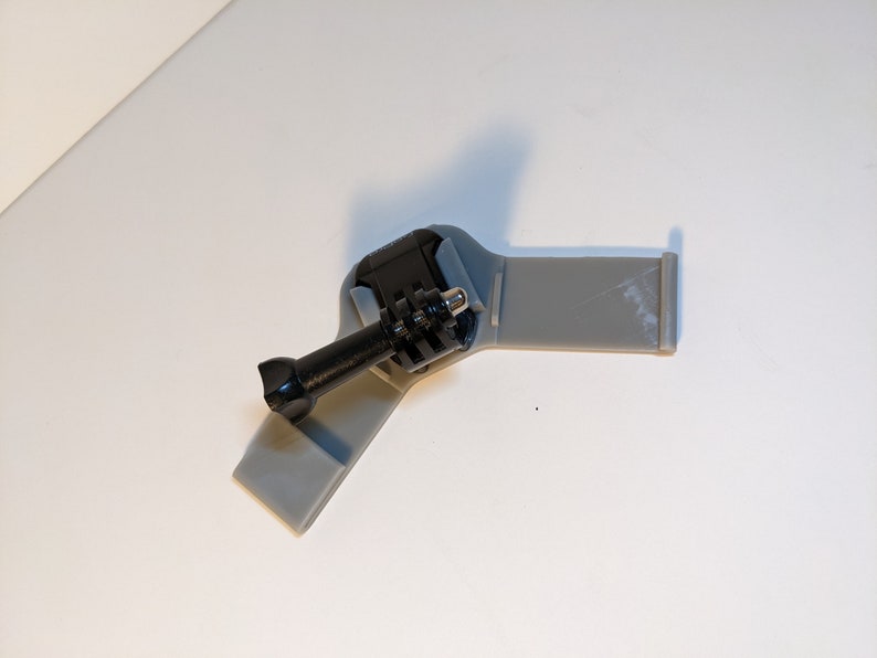 Pu&ograve; includere: Un supporto per fotocamera in plastica grigia con un supporto per fotocamera in stile GoPro nero attaccato.
