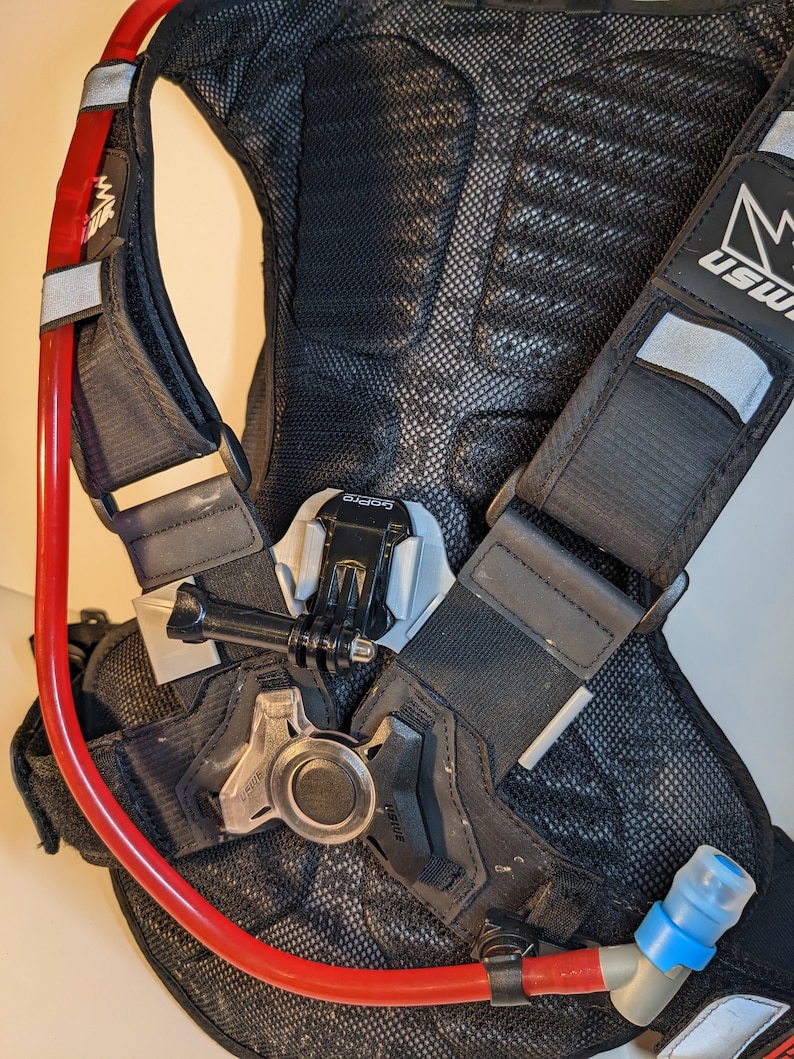 Pu&ograve; includere: Zaino di idratazione nero con un tubo di idratazione rosso e un beccuccio blu. Lo zaino ha un pannello posteriore in rete e un logo bianco e nero che dice "USWE". Lo zaino ha un supporto GoPro nero e argento.