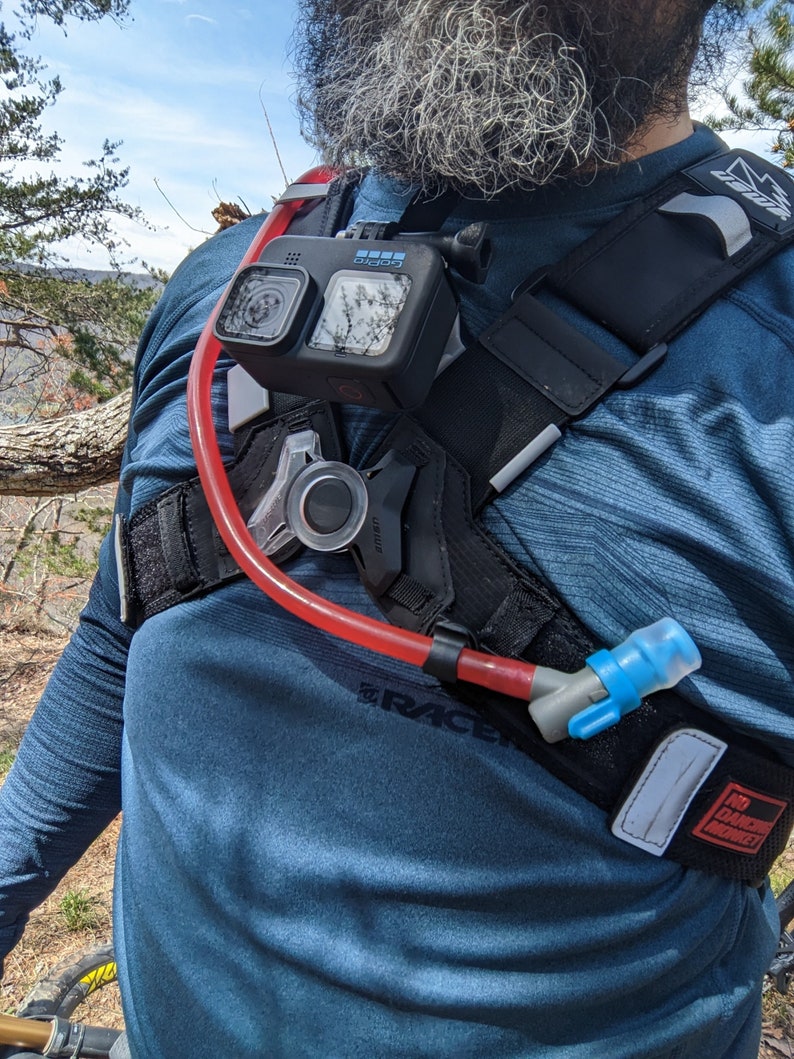 Pu&ograve; includere: Uno zaino di idratazione nero e rosso con una fotocamera GoPro montata sulla cinghia del petto. Lo zaino ha un tubo di idratazione blu con una valvola a morso. Lo zaino &egrave; indossato da una persona con la barba.