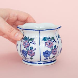 May include: A white ceramic planter with a scalloped rim, decorated with blue and purple floral patterns. The small, round planter has a blue base and trim. The planter is held by a hand against a pink background.